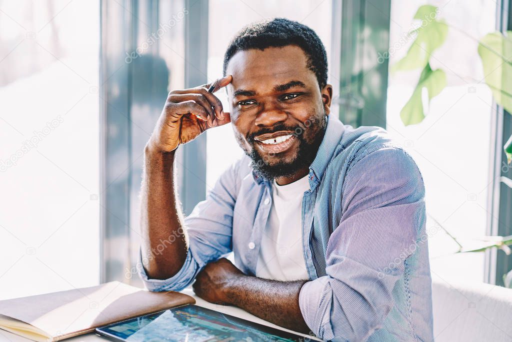 Portrait of cheerful african american hipster guy sitting at desktop smiling in coworking space, happy dark skinned blogger thinking about creative idea for article satisfied with working process 