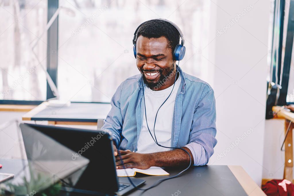 Smiling african american hipster guy in headphones connected to laptop computer learning by listening audiobook, positive dark skinned man entertaining with music in earphones working on freelance