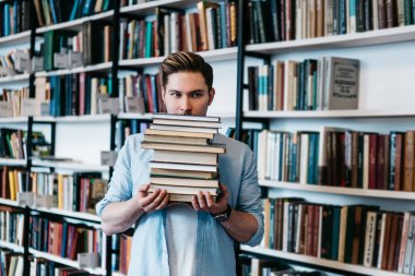 Rahat kıyafet çok edebiyat kitap bilim ders çalışma için elinde tutan overworked inatçı genç adam. Kütüphane iç kitap rafları ile ayakta ders kitapları ile akıllı öğrenci