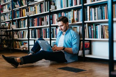 Modern dizüstü bilgisayarda genel kablosuz internet kitaplığında kullanarak Web sitesinde birçok edebiyat ve arama bilgi ile büyük bookshelf yakın katta oturan rahat kıyafet dalgın beyaz öğrenci