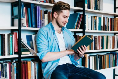 Concentrated smart hipster student reading textbook and preparing for studying seminar.Pensive young man holding literature book in hands sitting on chair near bookshelf in public library clipart