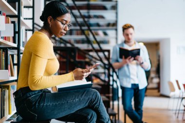 Kitaplığındaki sınavı için hazırlarken Smartphone cep telefonu ile gelen sms mesajý okuma dalgın Afrikalı-Amerikalı genç kadın. Öğrenciler boş zamanlarında üniversite kitabevi edebiyat kitap okuma harcama