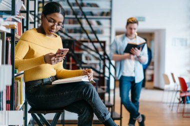 Afrikalı-Amerikalı Kitaplığı'nda oturan kablosuz Internet bağlantısı kullanarak modern smartphone cep telefonu ile internet sitesinde haber okumak. Hücresel hipster adam arka plan üzerinde kitap okuma sırasında güncelleştirme öğrenci koyu derili