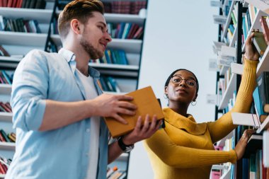 İki erkek ve kadın çok kültürlü hipsters görünümü altında modern Kütüphane içinde bookshelf kitaplardan çekerek formal giyim giymiş. Çeşitli gençlerin üniversite iç ders kitaplarının seçimi
