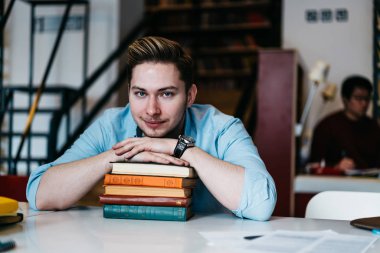 Yarım uzunluk portre yakışıklı beyaz öğrenci sınava hazırlık için pek çok kitap ile masaüstünde oturan kamera bakıyor. Genç adam rahat kıyafet Kitaplığı'nda eğitim vakit gülümseyen