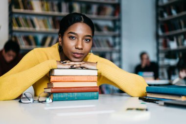 Kütüphanesi'nde masaüstü ile ders çalışma yazma ve ödev yapmak için birçok edebiyat ders kitapları içinde otururken kameraya bakıyor güzel Afrikalı-Amerikalı öğrenci portresi yarım uzunluk