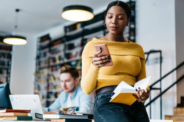 Afrikalı-Amerikalı genç kadın Kitaplığı'nda oturan elinde kitap tutarken gelen bildirim Smartphone cep telefonu ile okuma. Koyu tenli öğrenci hücresel kablosuz ortak Internet kullanarak posta kontrol