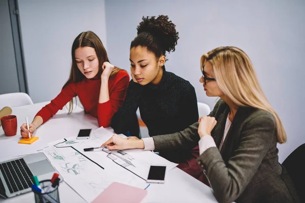 Skilled male teacher explaining new information for female students during lesson in college pointing on sketches, businesswoman having conversation with designers about plan and graphics on meeting