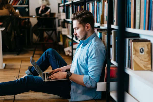 Side view of pensive hipster student searching information on internet websites for course work on laptop sitting near bookshelf in public library.Smart young man preparing for exam using computer