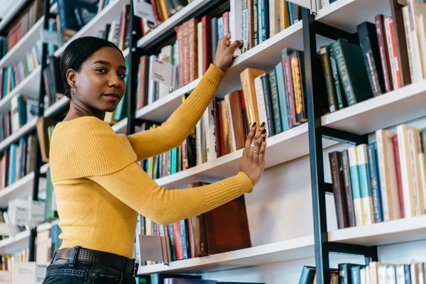 半长肖像的非洲裔美国年轻女子在休闲穿着看着相机 同时从书架上的公共图书馆拉文学书籍 深色皮肤的学生选择学习课本 — 图库照片