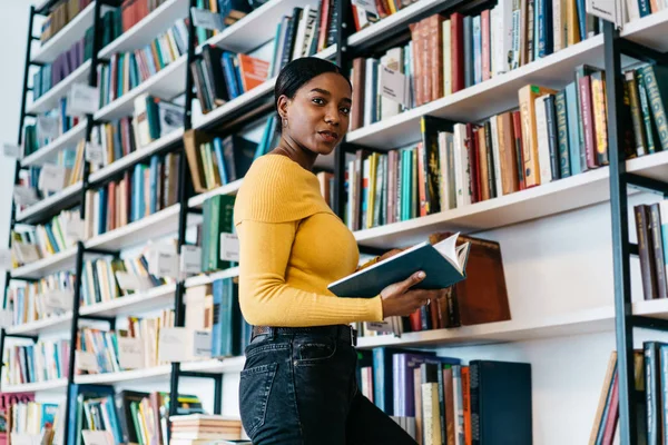 Portrait of positive african american young woman holding literature book in hands and smiling at camera.Dark skinned student preparing for writing course work searching information in textbook