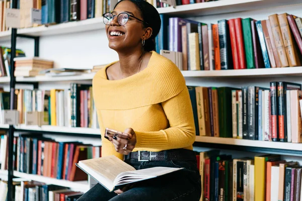积极的非洲裔美国年轻女子在时尚眼镜视觉矫正笑 而收到有趣的通知现代智能手机设备坐在现代图书馆的文学书籍 — 图库照片