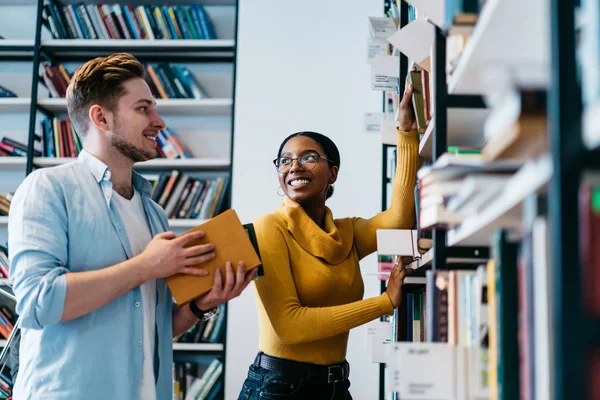 积极的两个多文化青年 Teamworking 在图书馆 开朗多样的时髦学生穿着正式的服装从书架上抽出书本选择文学写作课程工作 — 图库照片
