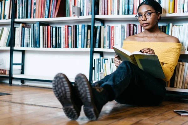 Portrait of attractive african american young woman looking at camera while sitting on floor near bookshelf and reading book.Charming smart dark skinned student with literature bestseller in library