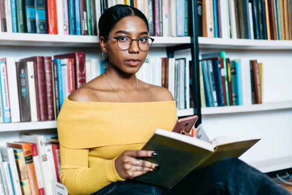 Portrait of dark skinned young woman in eyeglasses holding smartphone in hands and looking at camera while spending leisure time on reading literature book.African american student in library