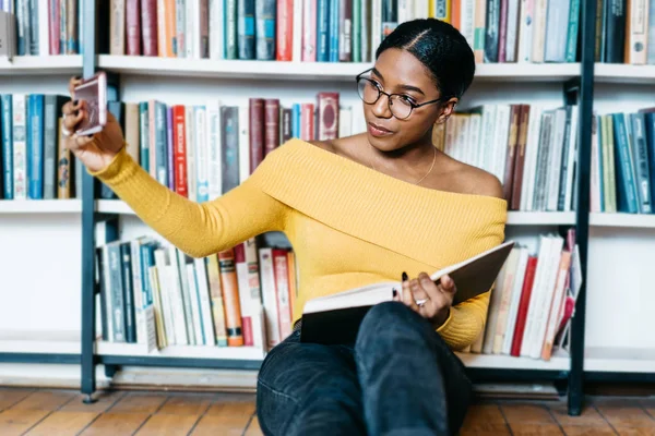 非洲裔美国青年女学生在眼镜制作自拍照片上的智能手机前相机共享网络坐在地板上的文学书籍和准备考试在图书馆 — 图库照片
