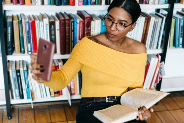 深色皮肤的女性博客 摆在智能手机前摄像头 并在图书馆与书自拍照片的社会网络 非洲裔美国学生在蜂窝上拍照 — 图库照片