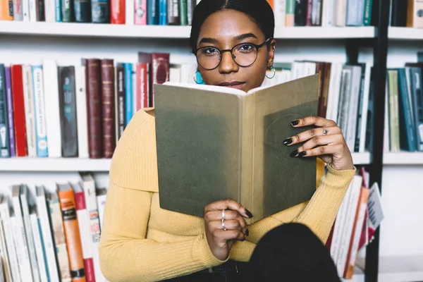 Half length portrait of attractive african american young woman in optical eyeglasses for vision correction looking at camera while holding literature book in hands sitting in modern library