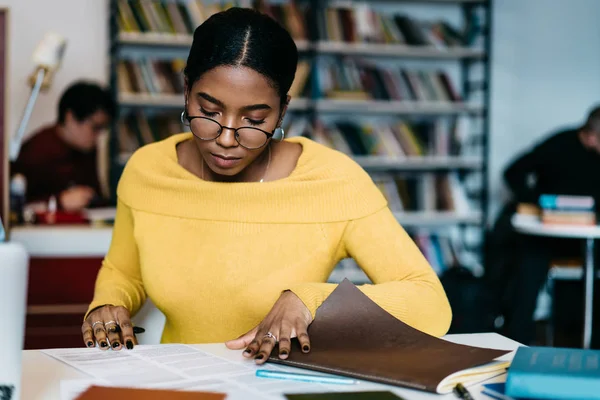在图书馆里沉思的聪明的非洲裔美国学生阅读学习任务和做作业 聚焦深色皮肤的年轻妇女在眼镜准备研讨会坐在桌面与抄写本旁 — 图库照片