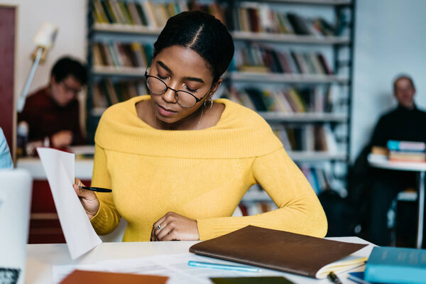 Concentrated african american hipster student in eyeglasses reading studying test and preparing for upcoming exams sitting at desktop in library.Pensive dark skinned young woman checking compendium