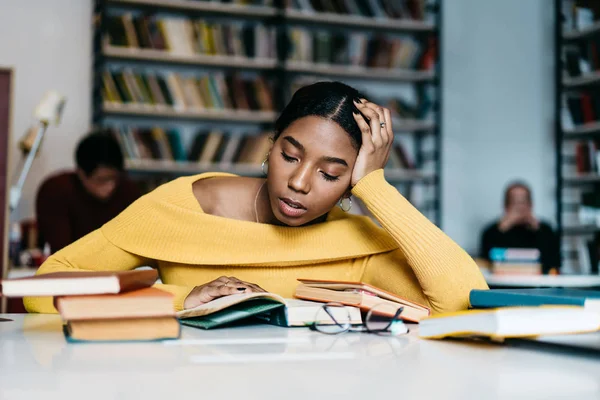 沉思的非洲裔美国年轻妇女阅读文学书籍准备写作课程的工作 智能深色皮肤的时髦学生在图书馆坐在桌上的教科书中搜索信息 — 图库照片