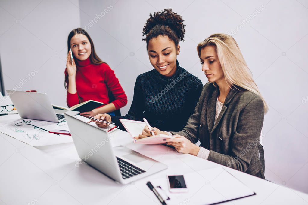 Serious multiracial women preparing report for startup searching information from different sources, working crew of female designers concentrated on their tasks using modern technology in office