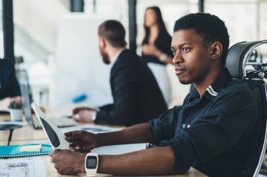 Dalgın Afrikalı-Amerikalı genç adam grafik kroki modern ofis iç tabloda toplantı oturan elinde tutan formal giyim giymiş. Koyu tenli yaratıcı erkek tasarımcı çizim ile