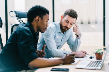 Resmi iki çok kültürlü meslektaşları problem çözme ve masadan ofiste dizüstü bilgisayar ile proje geliştirme teamworking giymek. Dalgın öğretmen ile birlikte koyu tenli öğrenci test yazma