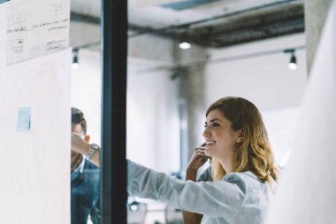 Planı üzerinde çalışması sırasında modern ofis meslektaşları ile işaret ederken gülüyor neşeli beyaz genç kadın. Kadın arabada pozitif formal giyim Atölyesi duran cam duvarın arkasında iletken