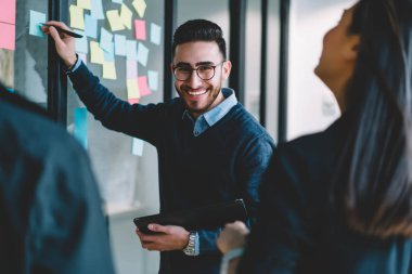 Eğitim kursları Office'te üzerinde meslektaşları ile işbirliği sürecinde duvara yapıştırılmış kağıt renkli çıkartmalar üzerinde notlar aşağı yazarken kameraya gülümseyen mutlu genç adam portresi yarım uzunluk
