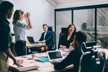 Çok kültürlü erkek ve kadın ofis çalışanları giyinmiş formal giyim eğleniyor finans şirketi çalışma yerinde oturuyor. Komik proje hakkında konuşurken gülüyor olumlu modern iş arkadaşları