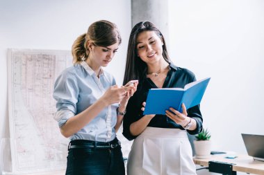 İki çok kültürlü olumlu genç kadın giyinmiş formal giyim ders kitabı okuma ve smartphone kullanarak bağlı 4 g için Internet ofis takım çalışması sırasında. Hippi kız bilgi telefon arama