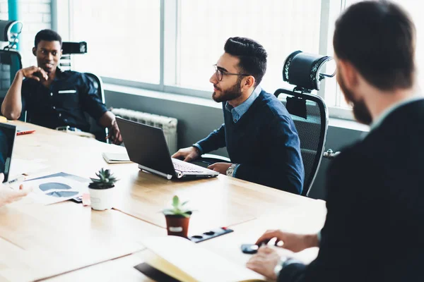 Diverse group of professionals dressed in formal wear communicating with each other and discussing business plan of developing productive strategy during briefing at meeting table in modern office