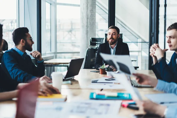 Male proud ceo talking with experienced employees and discussing ideas for successful project sitting at meeting table in modern office.Executive manager teamworking with creative young people