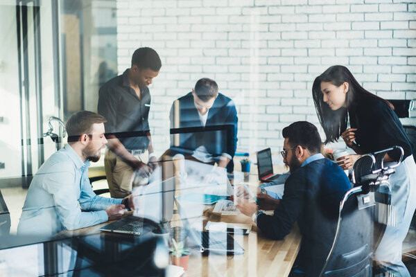 Diverse young people in formal wear communicating with each other and discussing creative ideas for developing design project sitting at meeting table behind glass wall in stylish office interior