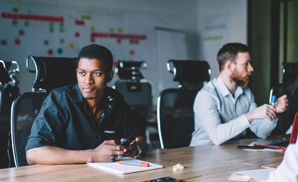 African american young man in formal wear updating profile on smartphone while sitting at meeting table and cooperating with colleagues on startup project in modern office interior