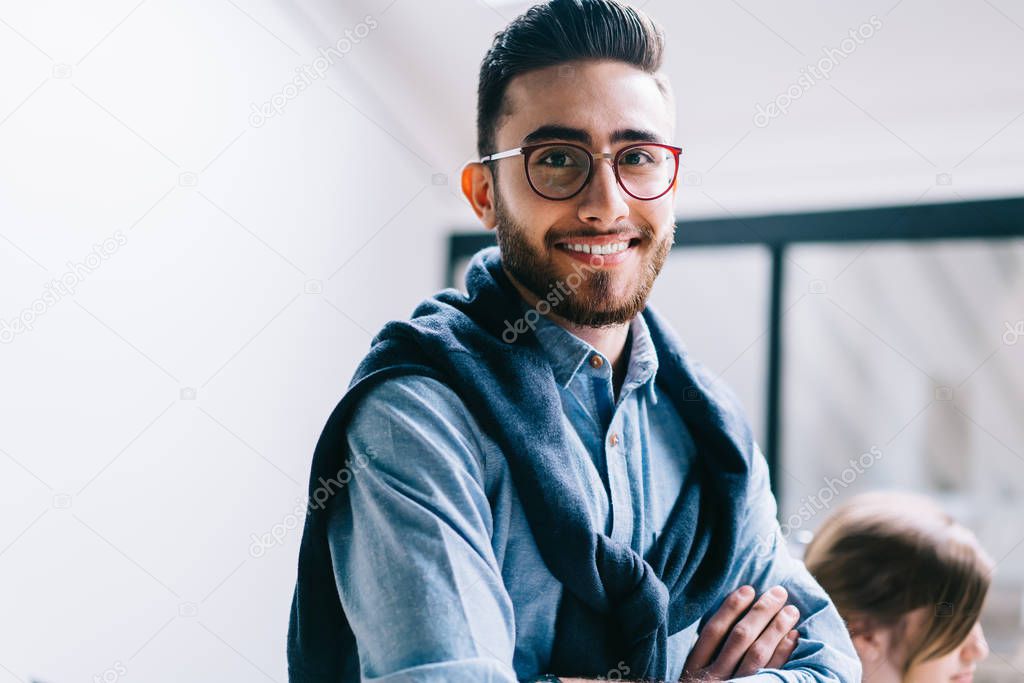Half length portrait of successful young entrepreneur in eyeglasses for vision correction smiling at camera.Positive businessman in formal wear and crossed hands standing in modern office