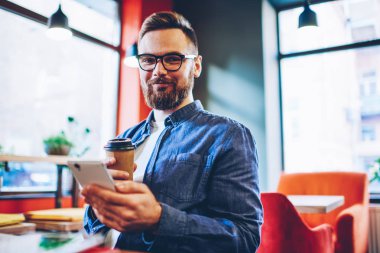 Yarım uzunluk portre başarılı sakallı erkek Blogger posta Smartphone cep telefonu ile 4 g ile sosyal ağlar denetlerken kameraya gülümseyen elinde lezzetli kahve ile internet. Cep ile pozitif adam