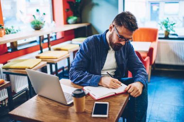 Dalgın yetenekli sakallı öğrenci Defteri'nde yer coworking yılında modern dizüstü bilgisayar oturma planı eğitim aşağı yazma kot gömlek giymiş. Not defterinde metin bilgileri belirterek hipster adam