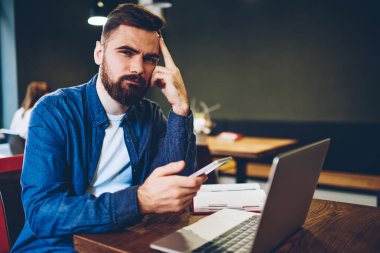 Yarım uzunluk portre ciddi hipster Blogger smartphone elinde tutan ve kameraya bakarken modern netbook oturan denim giyim giymiş. Kendine güvenen genç adam cep üzerinde App yükleme