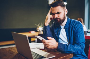 Konsantre sakallı genç işadamı bildirim istimal özgür 4 g Smartphone cep telefonu ile finans haberleri ile okuma Internet. Dizüstü bilgisayar başında otururken üzerinde hücresel mesajlaşma dalgın hipster blogger