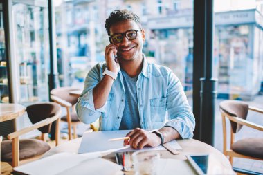 Başarılı yaratıcı erkek tasarımcı kameraya Studio yeni taslak üzerinde çalışma sırasında smartphone aygıt üzerinde konuşurken gülümseyen portresi. Neşeli hindu mimar Eyewear cep üzerinde arama