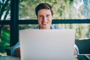 Modern Dizüstü aygıtı yüksek kullanarak web sayfalarında bilgi ararken kameraya gülümseyen pozitif genç adam portresi hızda kablosuz internet. Netbook çalışma başarılı erkek çevirmen