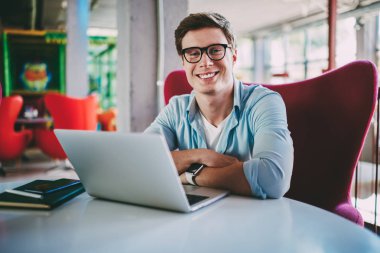 Modern kahve dükkanında kablosuz Internet bağlantısı kullanarak app yüklemek için dizüstü bilgisayar başında oturan ve yazılım güncelleme yaparken kameraya gülümseyen gözlük müreffeh genç adam portresi