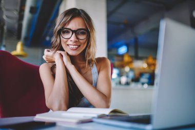 Uzaktan dizüstü cihaz çalışırken kameraya gülümseyen sevimli pozitif kadın serbest meslek portresi. Başarılı güzel öğrenci ders kitabı oturma ve netbook kafeteryasında sınav hazırlama