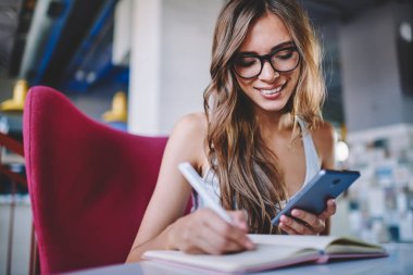 Masada oturan ve ücretsiz Wi-Fi bağlantısıyla smartphone üzerinde arama bilgi için Not Defteri'nde notlar yapma gözlük olumlu genç kadın. Neşeli öğrenci cellula bildirimini okuma
