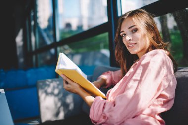 Kameraya eğitim kitap elinde tutan ve rahat coworking uzayda oturan yaklaşan sınavlar için hazırlanıyor süre seyir çekici genç kadın portresi. Akıllı kadın okuyucu ile en çok satanlar