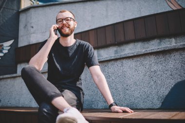 Gözlük smartphone aygıt üzerinde gezici arkadaşı ile iletişim sırasında kameraya gülümseyen sakallı neşeli genç adam portresi. Telefon aygıtı üzerinde arama olumlu hipster blogger
