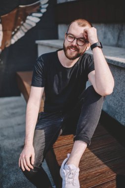 Pozitif sakallı genç adam modern şehir kentsel ortamda oturan rahat kıyafet giymiş. Neşeli hipster adam gülüyor ve uzağa sokakta boş zamanlarında dinlenme süre seyir şık gözlük