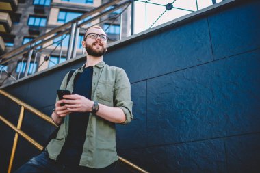 Aşağıda görünümü düşünceli erkek blogger düşünce modern smartphone kentsel ortamda merdivenlerde açık havada duran üzerinde internet yayıncılığı. Düşünceli hipster adam uzakta süre irsal sms arıyorsunuz